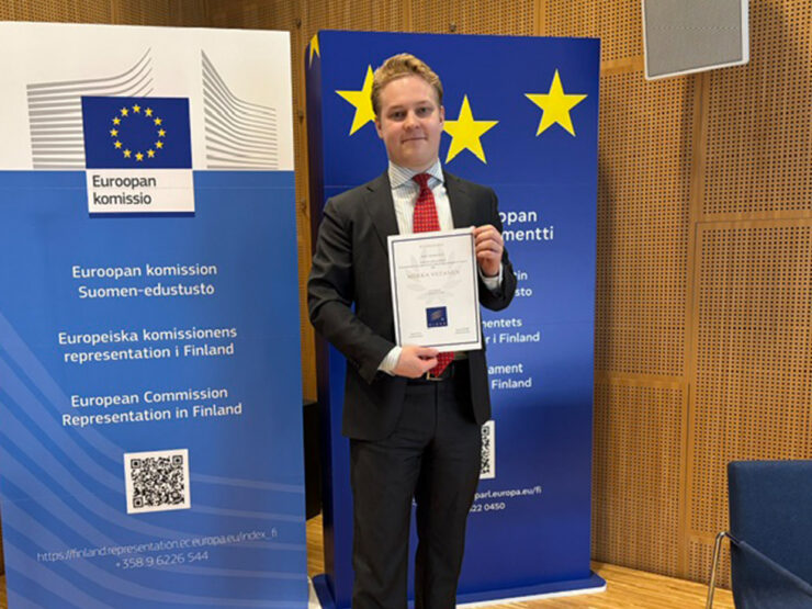 Miikka Viitanen pitää käsissään kunniakirjaa, taustallaan Euroopan komission ja Euroopan parlamentin rullajulisteet.