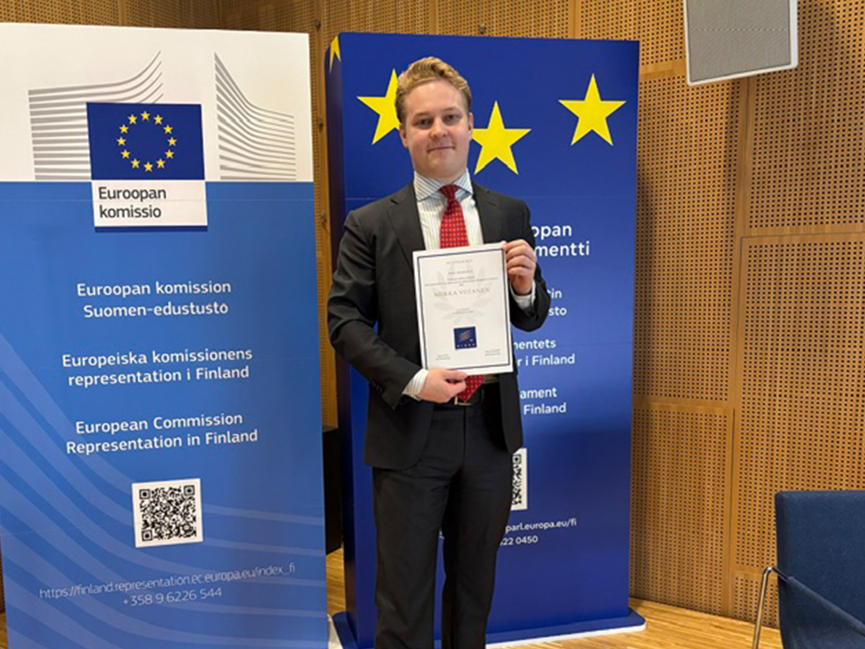 Miikka Viitanen pitää käsissään kunniakirjaa, taustallaan Euroopan komission ja Euroopan parlamentin rullajulisteet.