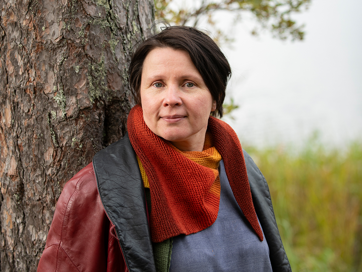 Maria Huhmarniemi seisoo selkä vasten mäntyä, taustalla kajastaa lehtipuun oksia, pitkää kasvillisuutta ja vettä.