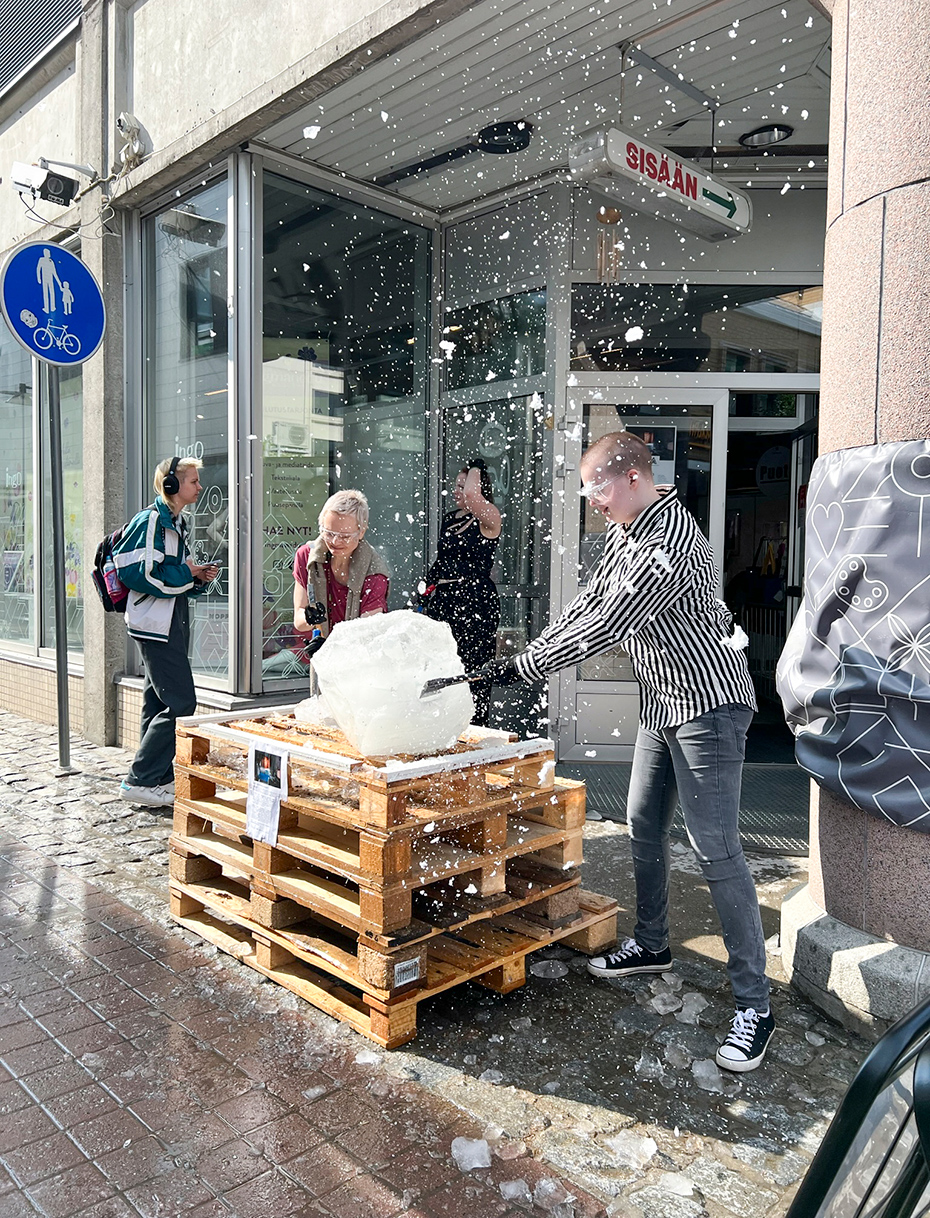 Kuva performanssista, jossa kaksi henkilöä lyö työvälineillä palasiksi isoa, kuormalavojen päällä olevaa jääkimpaletta keväisellä kadulla rakennuksen oven edessä ja ilmassa leijuu jäähiutaleita.