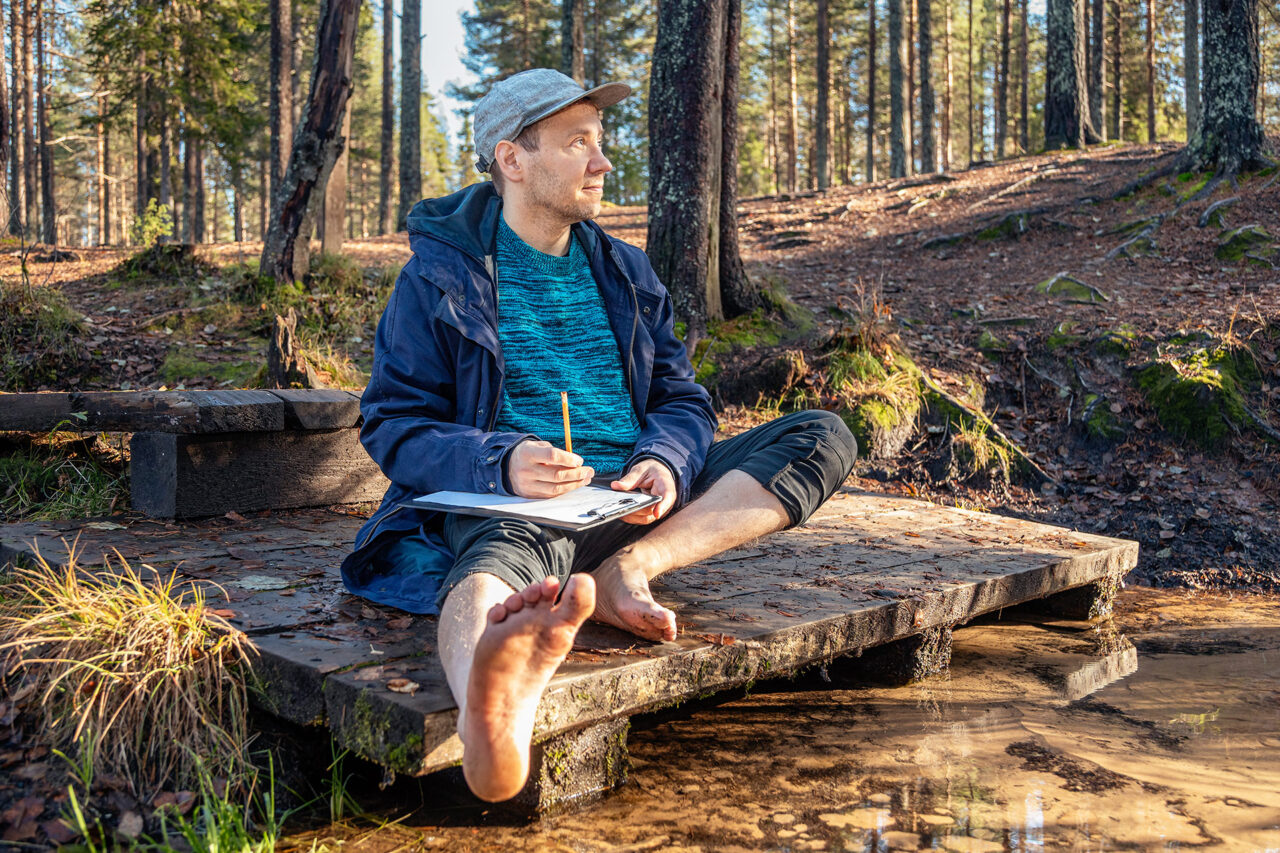 Jaakko Ruuska istuu paljain jaloin metsässä, lehtiö ja kynä kädessä.